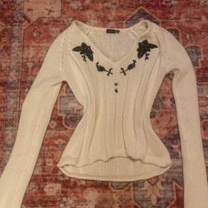 Vit stickad tröja med broderi från Vero Moda - Supersöt vit stickad tröja från Lindex med svarta broderade blommor vid v-ringningen. Tröjan har lång ärm, ribbad struktur och figurnära passform. Perfekt för lager-på-lager och snygg till både jeans och kjol.