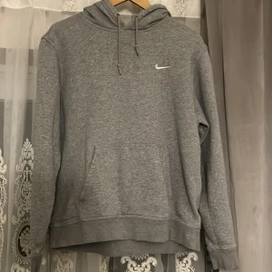 Grå Nike hoodie med huva - Säljer en grå hoodie från Nike med klassisk broderad swoosh på bröstet. Tröjan har huva med snörning och en stor magficka. Perfekt för chill dagar eller när du vill ha en enkel och stilren look.