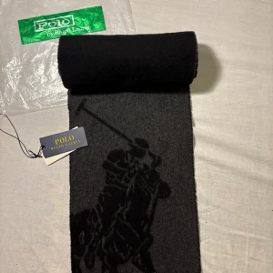 Svart och grå halsduk från Polo Ralph Lauren - Stilren halsduk från Polo Ralph Lauren i svart och mörkgrått med ikoniskt polospelare-motiv. Halsduken har fransar i ändarna och är tillverkad i mjuk ull, perfekt för kalla dagar. Klassisk design som lyfter vilken outfit som helst.