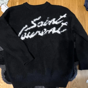 Svart stickad Saint Laurent tröja - Svart stickad tröja från Saint Laurent med vit logga i graffiti-stil över bröstet. Tröjan är helt oanvänd pga av storlek, skriv vid funderingar