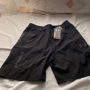 Svarta träningsshorts från Under Armour - Svarta Under Armour HeatGear shorts med elastisk midja och snörning. Lätt och luftig polyester, loose fit och coolt logotryck på benet. Snygga detaljer längs sidorna och diskret Armour-text. Perfekta för gymmet eller sport.