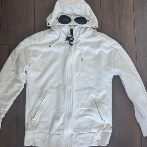 Vit hoodie med glasögon från C.P. Company - Säljer en vit hoodie från C.P. Company. Använd fåtal gånger. Och är i bra skick. Koftan är i storlek S.   (Fysiskt kvitto finns)