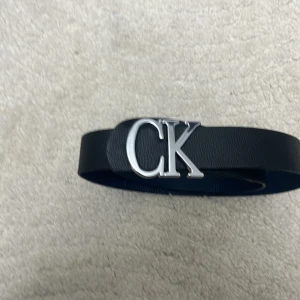 Svart skärp från Calvin Klein - Snyggt svart skärp från Calvin Klein med bred rem i skinn och en stor, blank silverspänne med CK-logga. Perfekt accessoar för att lyfta din outfit med en stilren och modern touch.