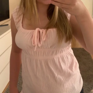 Ljusrosa linnetopp med knyt - Säljer en söt ljusrosa linnetopp med knyt framtill och rynkad struktur. Toppen har breda axelband och en mjuk, luftig känsla som passar perfekt till varma dagar. Supersnygg att matcha med jeans eller shorts för en chill look.