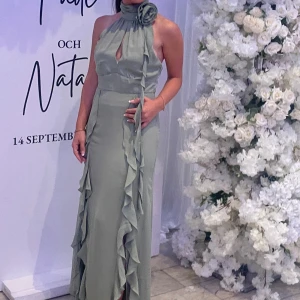 Ljusgrön långklänning med volang - Supersnygg ljusgrön långklänning med hög hals och stor blomdetalj vid halsen. Klänningen har ärmlös design, volanger längs framsidan och en liten slits. Perfekt för dig som vill sticka ut med en elegant och unik look.