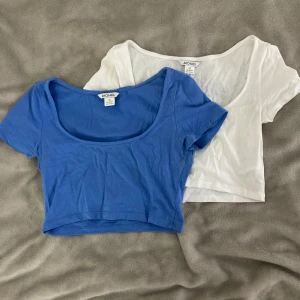 2 croppade toppar från Monki XS - Två croppade toppar från Monki i storlek XS. En är blå och en är vit, båda med rundad, djup urringning och korta ärmar. Perfekta att styla med höga jeans eller kjol. Tillverkade i mjuk bomull för en skön känsla.