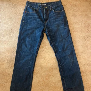 Blå loose fit jeans från Sir Benni Miles - Säljer ett par blå jeans från Sir Benni Miles med loose fit. Jeansen har snygga kontrastsömmar, klassiska fem fickor och unika vita detaljer på bakfickorna. Märkespatch i orange baktill och gylf med knapp. Perfekta för en avslappnad streetstil.