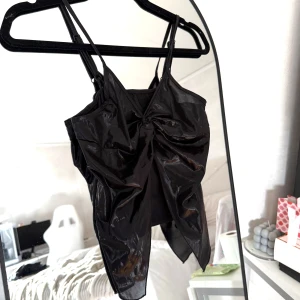 Svart glansig linnetopp - Svart glansig linnetopp i storlek S. Toppen har smala axelband, asymmetrisk nederkant och en snygg twistad detalj framtill. Materialet skimrande finish som ger en cool look.