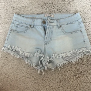 Ljusblå jeansshorts från Denim Life - Snygga ljusblå jeansshorts från Denim Life med fransig kant och klassisk femficksmodell. Shortsen har dragkedja, knapp och coola nitar bak i midjan. Perfekta för varma dagar och ger en avslappnad vibe.