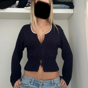 Mörkblå kofta med pärldetalj - Säljer en mörkblå, croppad kofta med långa ärmar och snygg pärldetalj längs den djupa ringningen. Koftan är stickad och har en figurnära passform som framhäver midjan. 