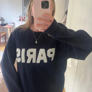 Mörkblå sweatshirt med PARIS-tryck - Cool mörkblå sweatshirt med stort vitt PARIS-tryck framtill. Tröjan har en klassisk rund halsringning och långa ärmar. Perfekt för dig som gillar streetstyle och vill ha en chill vibe. Mjuk och skön, passar till jeans eller leggings.
