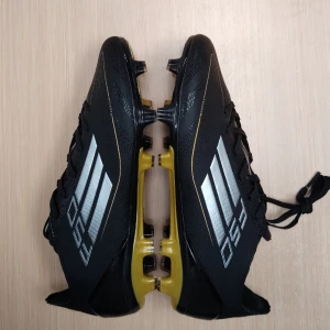 Adidas F50 Messi fotbollsskor svart/guld - Säljer ett par helt nya Adidas F50 Messi fotbollsskor i svart med silvriga ränder och guldiga detaljer. Skorna har snörning, platt sula med fasta dobbar och är tillverkade i syntetmaterial. Perfekta för dig som vill sticka ut på planen med stilrena och snabba skor.