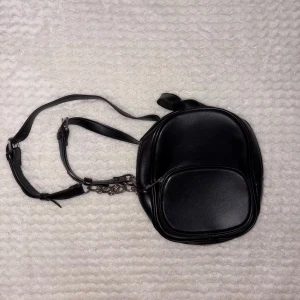 Svart miniryggsäck i skinnimitation - Snygg svart miniryggsäck med stilren design och justerbara axelremmar. Väskan har en rundad form, ett stort huvudfack och en mindre ficka framtill med dragkedja. Materialet är slätt och glansigt, och det finns en cool kedjedetalj på ena sidan.