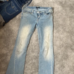 True Religion ljusblå bootcut jeans - Säljer ett par ikoniska ljusblå jeans från True Religion med bootcut passform. Jeansen har slitna detaljer framtill, klassiska kontrastsömmar och stora bakfickor med lock och silverfärgade knappar. Tillverkade i mjuk bomullsdenim för en skön känsla.