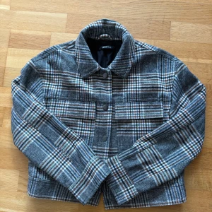 Rutig jacka från Gina Tricot - Snygg rutig jacka från Gina Tricot i grått, vitt,blått och brunt. Jackan har klassisk krage, knappar framtill och två stora bröstfickor. 