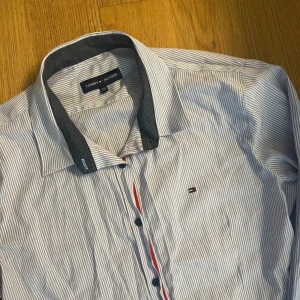 Randig skjorta från Tommy Hilfiger - STÅR I XXL MEN PASSAR M skjorta från Tommy Hilfiger med tunna blå och vita ränder, mörkare kontrastkrage och knappar framtill. Klassisk logga broderad på bröstet och coola manschetter med dubbla knappar. Perfekt för dig som gillar stilrena detaljer.