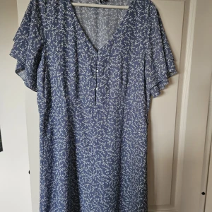 Blå blommig klänning Vero Moda XL - Söt blå klänning från Vero Moda i storlek XL med vitt blommönster. Klänningen har v-ringning, knappar framtill och korta, luftiga ärmar. Perfekt för en somrig look med sitt lätta tyg och avslappnade passform.