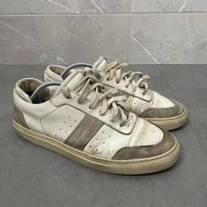  Axel Arigato skor - Snygga sneakers från Axel Arigato i vitt och beige med diskreta gulddetaljer. Skorna har klassisk låg profil, snörning och detaljer i mocka och skinn. Perforerade sidor och robust sula ger en clean och stilren look som passar till det mesta.