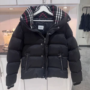Svart pufferjacka från Burberry - Säljer en svart pufferjacka från Burberry med klassiskt rutigt foder i huvan. Jackan har dragkedja och knappar framtill, två fickor med dragkedja och en oversized passform. Perfekt för kalla dagar. Aldrig använd!