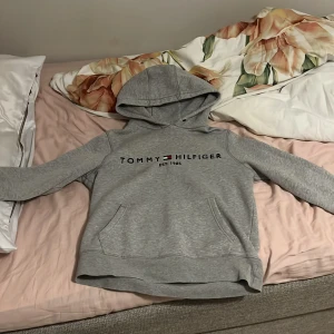 Grå hoodie från Tommy Hilfiger - Säljer en grå hoodie från Tommy Hilfiger med broderad logga och text på bröstet. Tröjan har huva, magficka och är fodrad med mjukt material på insidan. Perfekt för kyliga dagar och riktigt skön att ha på sig. Priset kan diskuteras passar även på folk som har storlek M