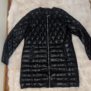 Svart quiltad lång dunjacka Amisu - Svart lång dunjacka från Amisu med snyggt quiltat mönster upptill och horisontella sömmar nedtill. Jackan har rund halsringning, dragkedja framtill och två fickor med dragkedja. Perfekt för kyliga dagar och ger en clean look.