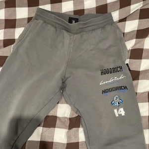 Grå mjukisbyxor från Hoodrich S - Grå mjukisbyxor från Hoodrich med tryck och broderade detaljer på benen och bakfickan. Byxorna har snörning i midjan, sidofickor och en bakficka. Perfekta för chill dagar och streetwear-stil.