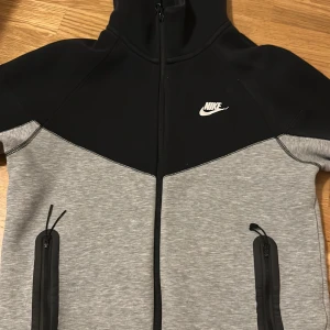 Nike Tech Fleece hoodie svart/grå - Nike Tech Fleece hoodie i svart och ljusgrått med hel dragkedja och två fickor med dragkedja. Klassisk Nike-logga på bröstet och huva. Perfekt för chill eller träning, riktigt snygg och sportig look.
