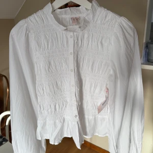 Vit smockad blus med volang från Nelly - Helt ny Supersöt vit blus från Nelly med smockad front och rygg, volangkant nertill och långa puffiga ärmar. Knappar hela vägen framtill och lätt rynkad struktur. Prislapp sitter kvar och är i storlek 34