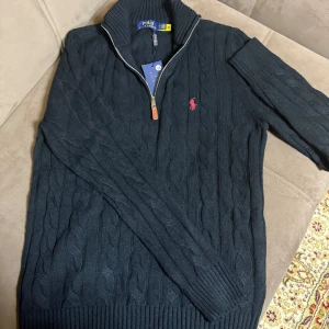 Svart stickad tröja Polo Ralph Lauren - Kabelstickad svart tröja från Polo Ralph Lauren med halv dragkedja och hög krage. Klassisk röd logga broderad på bröstet. Tröjan är långärmad och har ribbade muddar och nederkant. Perfekt för dig som gillar stilren och tidlös design. Passade inte mig i storleken då jag behövde L annars va den jättefin och stilig.