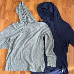 2 st hoodies från Polo Ralph Lauren M - Två snygga hoodies från Polo Ralph Lauren i storlek M. En är ljusgrå med liten broderad logga vid höften och den andra är mörkblå med röd logga på bröstet. Båda har huva med snörning och är tillverkade i mjuk bomull. Perfekta för chill dagar eller när du vill vara casual.