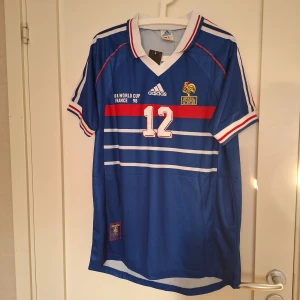 Frankrike VM 98 Henry #12 Adidas tröja - Klassisk blå fotbollströja från Adidas, Frankrikes VM 1998 med Henry #12 på ryggen. Tröjan har vita och röda detaljer, broderat tuppemblem och matchdatum mot Brasilien. V-ringad krage, kortärmad och tillverkad i lätt polyester. Perfekt för dig som älskar retro och fotbollshistoria.
