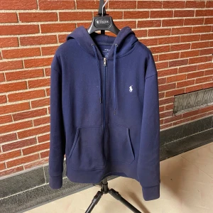 Mörkblå hoodie från Polo Ralph Lauren - Mörkblå hoodie från Polo Ralph Lauren med dragkedja, huva och snörning. Klassisk logga broderad på bröstet och två stora fickor framtill. Perfekt för dig som gillar en clean och stilren look.