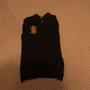 Svart pikétröja från Ralph Lauren - Size M. Snygg svart pikétröja från Ralph Lauren med klassisk krage och korta ärmar. På bröstet finns ett broderat emblem med nummer 3 och Polo-logga samt den ikoniska polospelaren i gult. Perfekt för en stilren och sportig look.