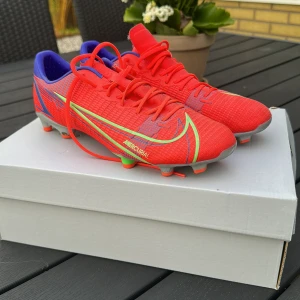 Nike Mercurial fotbollsskor röd/grön - Säljer ett par Nike Mercurial fotbollsskor i en riktigt snygg röd färg med gröna och blåa detaljer. Skorna har snörning, platt sula med dobbar och är tillverkade i syntetmaterial. Perfekta för dig som vill sticka ut på planen med stilren design och grym passform. Pris kan diskuteras vid snabb och smidig affär.
