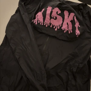 Svart vindjacka från Risky med tryck - Svart vindjacka från Risky i 100% nylon med dragkedja framtill. Stor rosa text med drip-effekt på ryggen ger jackan en edgy vibe. Perfekt för dig som gillar streetstyle och vill sticka ut med coola detaljer.