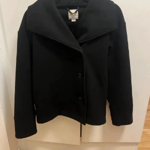 Svart kappa - Snygg svart kappa med knappar och bred krage. Perfekt för kyligare dagar och ger en stilren look. PRIS KAN DISKUTERAS!!!