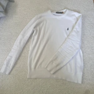 Polo Ralph Lauren tröja - Vit stickad tröja från Polo Ralph Lauren med klassisk rund halsringning och broderad logga på bröstet. Tröjan har ribbade muddar vid ärmslut och nederkant, perfekt för en clean och stilren look.