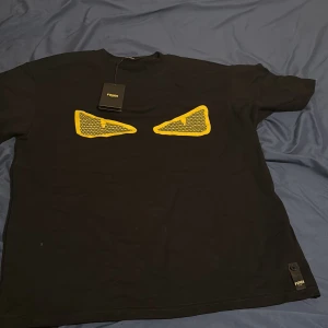 Svart Fendi t-shirt med gula ögon - Svart t-shirt från Fendi med ikoniska gula ögon framtill. Klassisk passform, rund hals och korta ärmar. Tillverkad i mjuk bomull och har Fendi-logga med knappdetalj nertill. Perfekt för dig som vill sticka ut med en lyxig streetstyle.