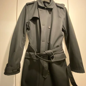 Svart trenchcoat från Topman, S - Stilren svart trenchcoat från Topman i storlek S. Jackan har klassisk dubbelknäppning, axelklaffar, bälte i midjan och knappar vid ärmsluten. Perfekt för dig som vill ha en clean och tidlös look. Materialet är mjukt och följsamt.