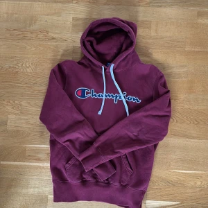 Vinröd hoodie från Champion - Säljer en vinröd hoodie från Champion med stor logga broderad på bröstet. Tröjan har huva med vita snören, känguruficka och är långärmad. Perfekt för dig som gillar en sportig och avslappnad stil.