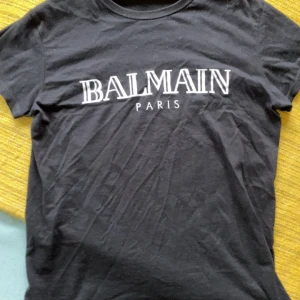Svart t-shirt med Balmain-tryck - Svart t-shirt i bomull med vit Balmain Paris-logga framtill. Klassisk rund hals och korta ärmar. Enkel och stilren modell som passar perfekt till jeans. Skön passform och mjukt material.