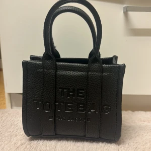 Svart The Tote Bag från Marc Jacobs - Säljer en svart The Tote Bag från Marc Jacobs i stilrent och tåligt skinn. Väskan har präglad logga framtill, två rejäla handtag och en boxig, strukturerad form. Perfekt för dig som vill ha en ikonisk och trendig handväska med plats för det viktigaste.