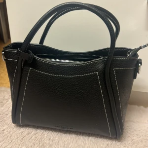 Svart handväska med vita sömmar - Snygg svart handväska  i skinn med vita kontrastsömmar. Väskan har två handtag, silverfärgade metalldetaljer och dragkedja upptill. Perfekt storlek för det viktigaste och stilren design som passar till många outfits.