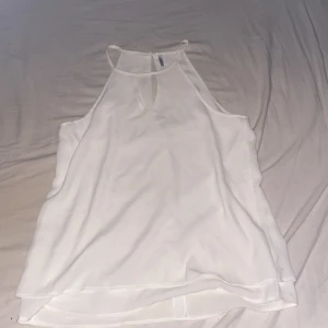 Vit ärmlös linnetopp med nyckelhålsdetalj - Säljer en stilren vit linnetopp med smala axelband och en liten nyckelhålsöppning framtill. Toppen är ärmlös och har en luftig passform, perfekt för varma dagar. Enkel och fräsch design som är lätt att matcha med olika outfits.