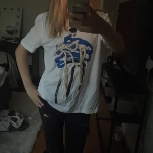 Vit t-shirt med blått tryck och snörning - Cool vit t-shirt med ett stort blått tryck framtill och unika snörningsdetaljer i beige som hänger ner. Modellen är kortärmad och har en loose passform, perfekt för dig som gillar statement-plagg med kreativ design.
