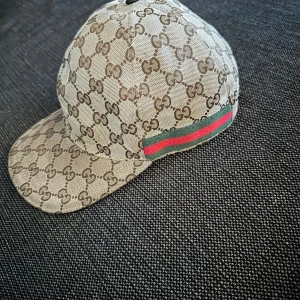 Gucci beige keps med GG-mönster - Snygg beige keps från Gucci med klassiskt GG-monogram i brunt över hela kepsen. På sidan finns den ikoniska gröna och röda randen. Kepsen har böjd skärm och insida i mörkbrunt tyg. Perfekt för dig som vill ha en lyxig och trendig accessoar.