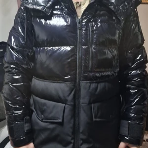 Svart pufferjacka Moose Knuckles - Snygg svart pufferjacka från Moose Knuckles med glansig finish och stora fickor framtill. Jackan har dragkedja, huva och är vadderad med REPREVE-material. Perfekt för kalla dagar och har en modern, urban look. Ny pris ca 8000. Kvitto finns. 