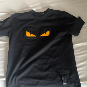Svart Fendi t-shirt med gula ögon - Svart t-shirt från Fendi i 100% bomull med ikoniska gula monsterögon och Fendi-logga på bröstet. Klassisk rund hals och korta ärmar. Perfekt för dig som vill sticka ut med en lyxig streetstyle-look.