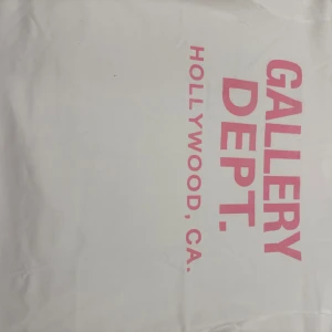 Vit t-shirt Gallery Dept. L - Vit t-shirt från Gallery Dept. med tryck i rosa på bröstet och texten 'HOLLYWOOD, CA.' på sidan. Klassisk rund hals och rak passform. Tillverkad i mjuk bomull, perfekt för en clean och stilren look.