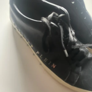 Svarta Philipp Plein sneakers - Snygga svarta sneakers från Philipp Plein med vita sulor och coola silverdetaljer längs sidan. Skorna har snörning och är tillverkade i skinn, med märkets logga tryckt på sidan. Perfekta för dig som gillar streetstyle och exklusiva märken.
